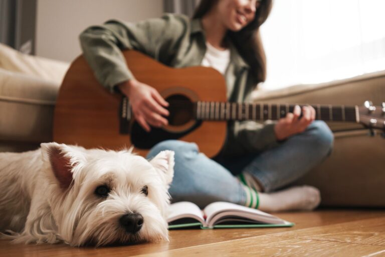 Happy white woman playing guitar while sitting on floor Música relaxante para cães: dona toca guitarra sentada no chão da sala com o seu cão ao lado