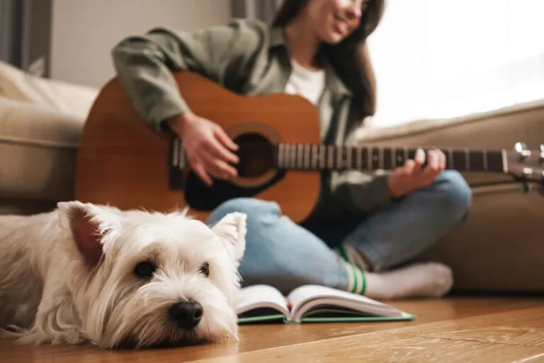 Happy white woman playing guitar while sitting on floor Música relaxante para cães: dona toca guitarra sentada no chão da sala com o seu cão ao lado